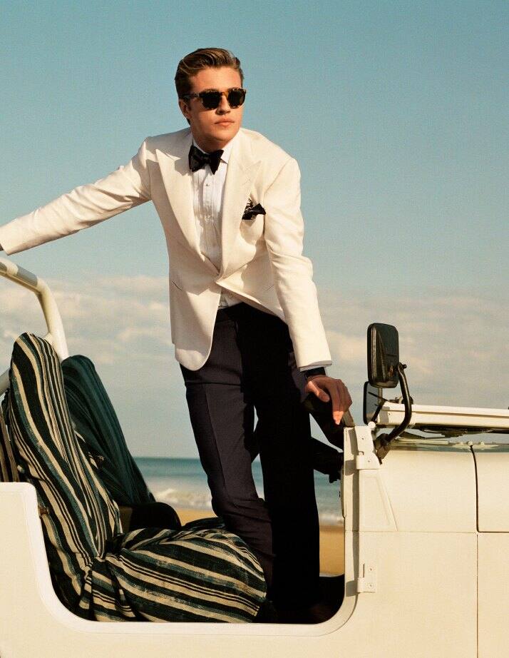 Man standing in Jeep wears white tuxedo jacket and black trousers.
